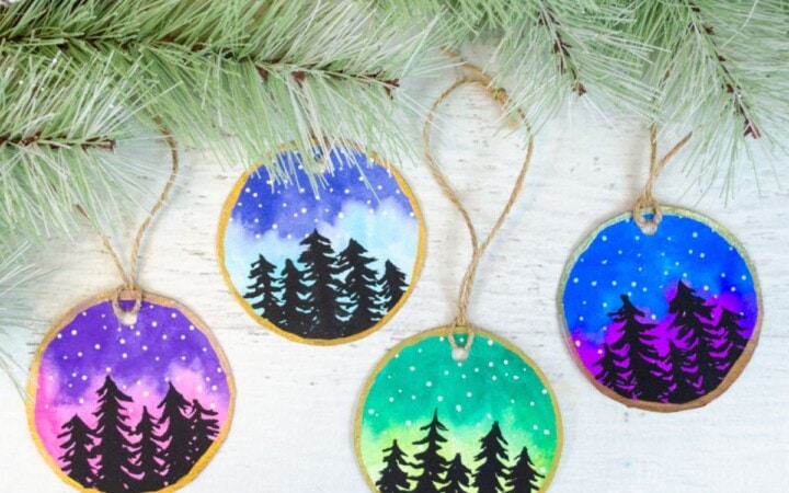 Watercolor Christmas ornaments with colorful sky and black trees with stars.