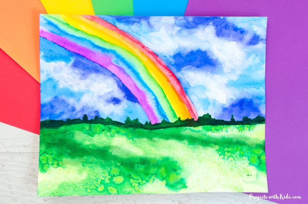 Rainbow art project for kids and tweens using watercolor paint. Bright rainbow in a stormy blue sky with grass. 