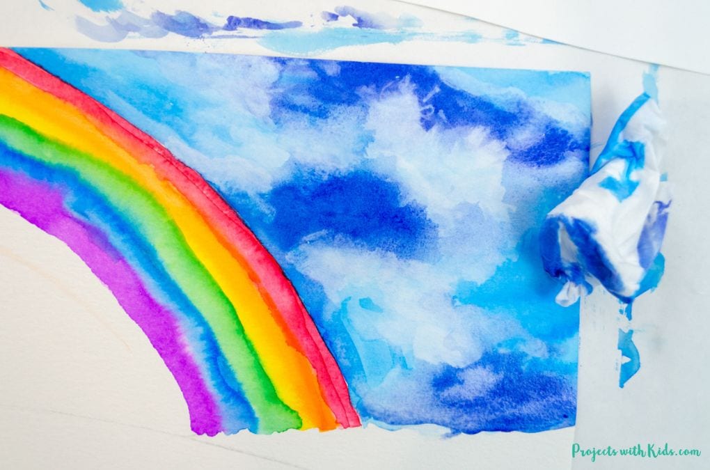 Using blue watercolor paint and tissue to paint clouds in a sky with a rainbow. Art project for kids and tweens.